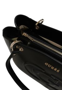 Schwarze Lederhandtasche mit goldenen Beschlägen, die über doppelte Oberöffnungen, ein internes Reißverschlussfach und ein geprägtes Logo auf der Vorderseite verfügt.
