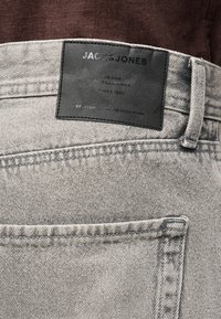 Ανοιχτό γκρι τζιν παντελόνι με μαύρη δερμάτινη ετικέτα Jack & Jones ραμμένη πάνω από την πίσω τσέπη, φορεμένο με καφέ πουκάμισο.