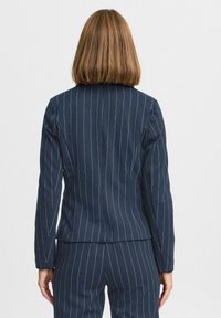 Kobieta z jasnobrązowymi włosami do ramion, ubrana w granatowy garnitur w prążki, stojąca tyłem na białym tle.