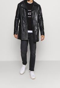 Manteau en cuir noir avec un large col, boutons croisés, porté sur un pull noir et un jean foncé, assorti à des baskets blanches.