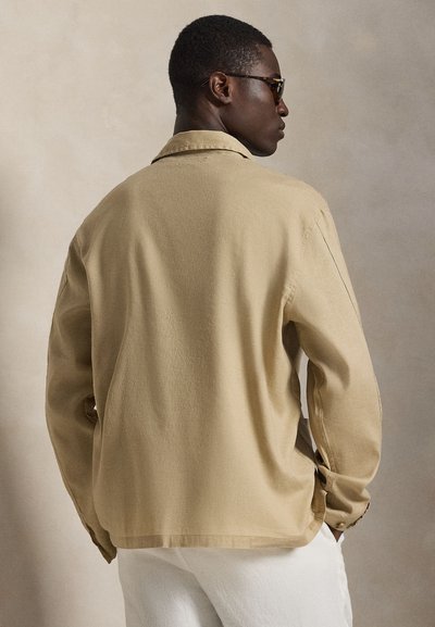 Άνδρας με μπεζ μπουφάν και άσπρο παντελόνι, εμφανίζεται από την πίσω πλευρά, στέκεται μπροστά σε ένα απλό, ουδέτερο φόντο.