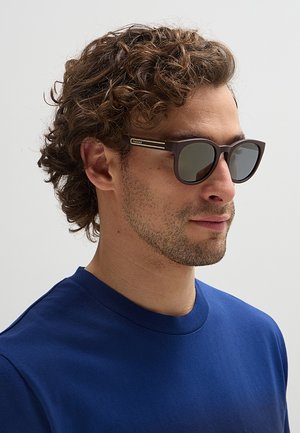 Uomo con capelli castani ricci che indossa occhiali da sole scuri e una maglietta blu a girocollo, rivolto verso destra su uno sfondo semplice.