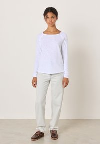 Junge Frau steht und trägt ein weißes Langarmshirt, helle Jeans und braune Schnürschuhe vor einem schlichten Hintergrund.