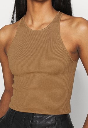 Close-up van een persoon die een mouwloos, geribbeld bruin tanktop draagt en zwarte leren broek, met lang donker haar zichtbaar.