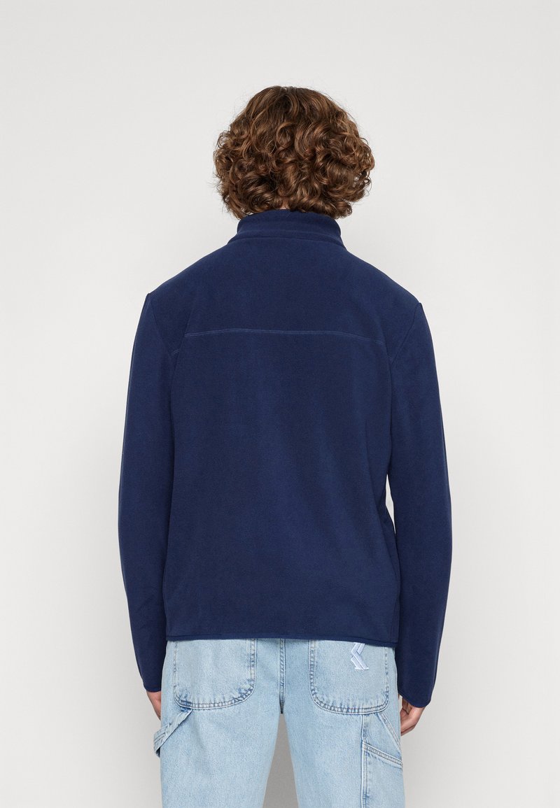 Granatowa polarowa kurtka z wysokim kołnierzem, gładką fakturą i prostym krojem. Posiada jeden szew na plecach.