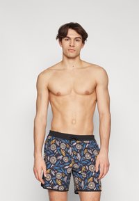 Shorts de bain fleuris avec une taille noire, présentant un motif botanique bleu, jaune et blanc sur un fond sombre.