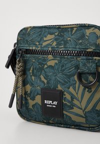 Borsa a fiori nei toni del verde scuro e oliva. Realizzata in tessuto resistente con chiusura a zip, presenta una patch con logo e cordini decorativi.