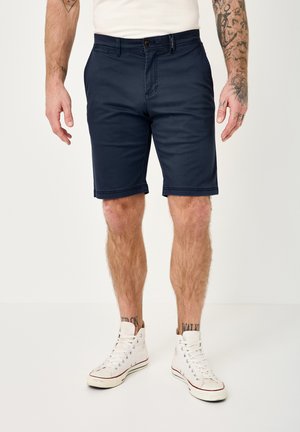 Man wearing navy blue knee-length shorts, white high-top sneakers, and a white t-shirt, standing with visible arm and leg tattoos.