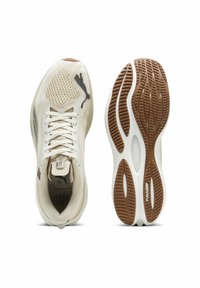 Idrottsskor med en beige meshöverdel, grå detaljer och en mönstrad gummisula med "PUMAGRIP"-logotypen. Snörad design, låg silhuett.