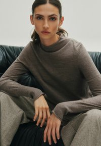 Brązowy sweter z dzianiny z wysokim kołnierzem i długimi rękawami, połączony z jasnoszarymi spodniami w wzór rybiej ości. Widoczne szczegóły teksturowej tkaniny.