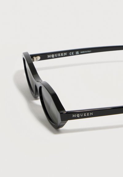 Μαύρα οβάλ γυαλιά ηλίου με χοντρά πλαίσια, λογότυπο "McQueen" στους βραχίονες και σκούρους φακούς, ακουμπισμένα σε λευκή επιφάνεια.