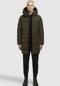 Olivno zelena podložena jakna s kapuco, z zadrgo spredaj, dvema stranskima žepoma in dolgim krojem. Obuta preko črnega oblačila in čevljev.