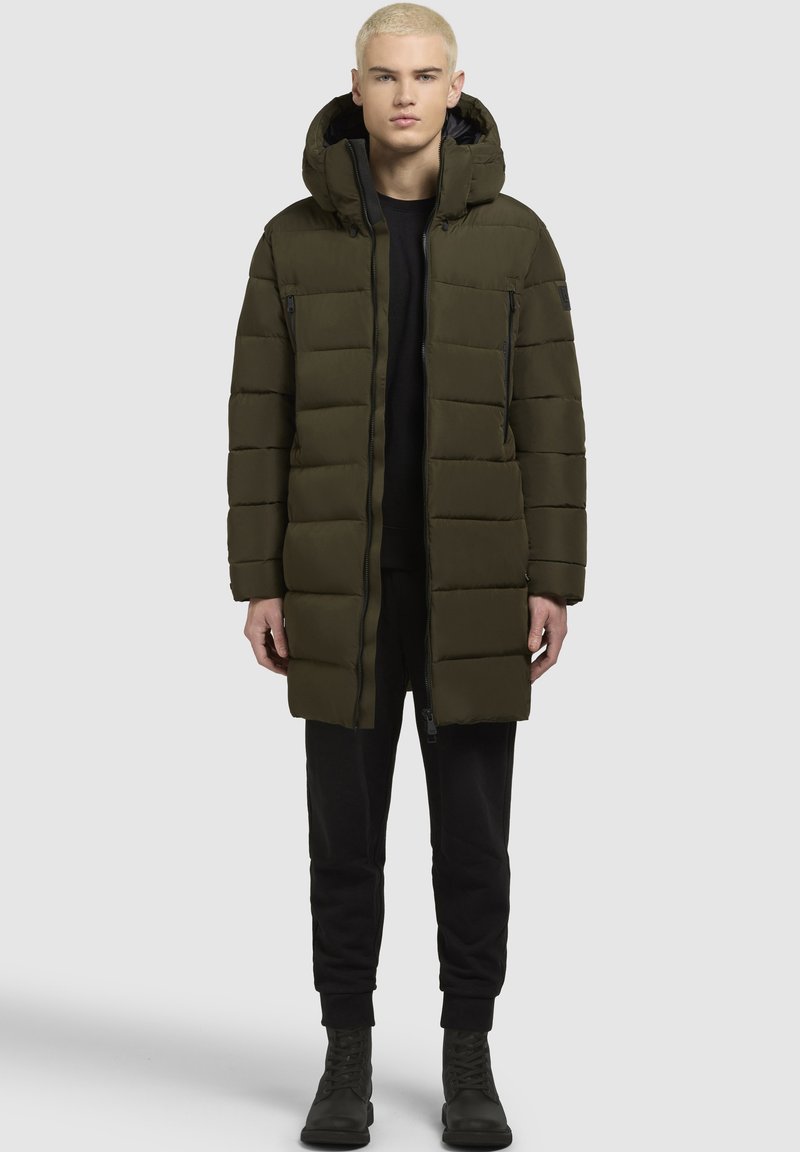 Olivno zelena podložena jakna s kapuco, z zadrgo spredaj, dvema stranskima žepoma in dolgim krojem. Obuta preko črnega oblačila in čevljev.