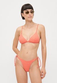 Bikini corail à côtes avec des bretelles fines et réglables et un bas à nouer. Le tissu a une finition texturée, mettant en valeur le design du maillot de bain.