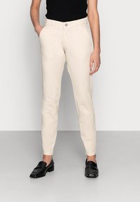 Calças bege sob medida com um corte ajustado, bolsos frontais e fecho em botão prateado, combinadas com sapatilhas pretas sobre um fundo cinza.