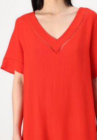 Vestido vermelho de mangas curtas com decote em V, apresentando um tecido texturizado e detalhes em rendas ao longo do decote e das mangas.