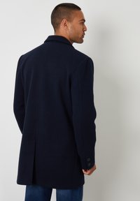 Marineblauer Wollmantel mit Reverskragen, Rücken mit einfacher Belüftung und zwei-Knopf-Manschetten, kombiniert mit blauer Jeans. Glatte Textur.