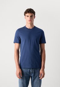 Hombre con una camiseta de mangas cortas azul marino lisa y jeans de color azul claro, de pie contra un fondo blanco liso.