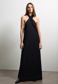 Maxi dress nera con allacciatura a halter, caratterizzata da una texture arricciata, silhouette ad A e design sobrio. Indossato con semplici calzature nere.