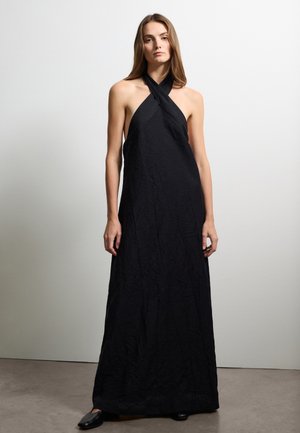 Schwarzes Maxib Kleid mit Neckholder, gekräuselter Textur, A-Linien-Silhouette und zurückhaltendem Design. Getragen mit einfachen schwarzen Schuhen.