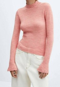 Woman wearing a fitted pink knit sweater with flared cuffs and ruffle details paired with high-waisted off-white pants.