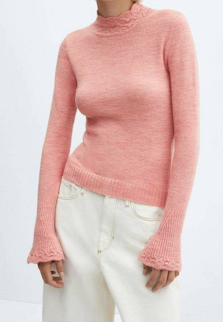 Woman wearing a fitted pink knit sweater with flared cuffs and ruffle details paired with high-waisted off-white pants.