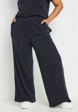Pantaloni a gamba larga blu navy con texture a righe verticali, dotati di tasche laterali e vestibilità comoda, abbinati a calzature bianche.