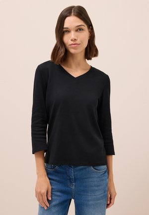 Schwarzes, langärmliges Oberteil mit V-Ausschnitt und lockerer Passform, aus einem weichen Stoff gefertigt. Kombiniert mit blauen Jeans.