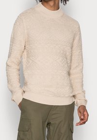 Beige stickad tröja med hög krage, med texturerade mönster och ribbad nederdel och ärmar, i kombination med olivgröna byxor.