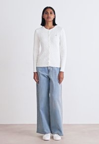 Witte kabelgebreide cardigan met een ronde halslijn, knoopsluiting, gecombineerd met lichtblauwe jeans met brede pijpen en witte sneakers.