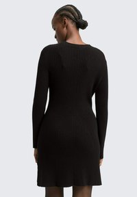 Schwarzes, geripptes Strickkleid mit langen Ärmeln, einer körpernahen Passform und ausgestelltem Saum. Die Textur ist ausgeprägt und zeigt vertikale Linien.