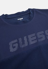 Námořnická modrá sportovní tričko s texturovaným síťovým vrškem, se výrazným bílým logem "GUESS" ve formě teček přes hruď a pevným spodním dílem.