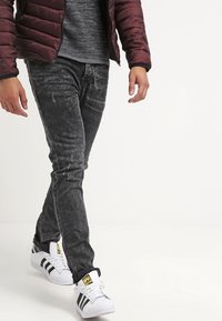 Jean noir avec un aspect délavé et usé, associés à des baskets blanches avec des rayures noires et un logo jaune. Les mains sont dans les poches.