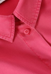 Jasnoróżowa koszula z kołnierzykiem o gładkiej teksturze, z dwoma guzikami oraz widocznymi detalami ściegów wzdłuż kołnierza i listwy guzikowej.