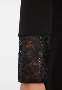 Schwarzes Kleidermodell mit einem soliden, glatten Stoff und einem Spitzenmanschette, die florale Muster aufweist und eine transparente Textur am Handgelenk hat.