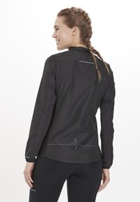 Sort letvægtsjakke med lange ærmer, der har reflekterende accenter og et bølge-mønster langs kanten. Glat tekstur og tætsiddende design.