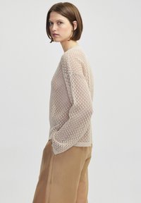 Maglione a maglia in beige chiaro con un tessuto largo e aperto e maniche lunghe, abbinato a pantaloni color sabbia. Presenta un collo rotondo e polsini a coste.