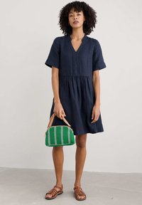 Navy linnen korte mouwen jurk met V-hals en gathers op de taille, gecombineerd met een groen-wit gestreepte handtas en bruine sandaaltjes met bandjes.