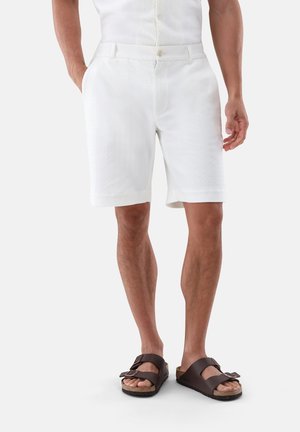 Mann trägt weiße, strukturierte Shorts und dunkelbraune Sandalen mit Doppelriemen und steht mit einer Hand in der Tasche.