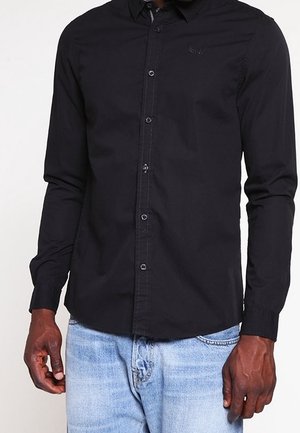 Man draagt een nauwsluitend zwart overhemd met knopen en lange mouwen, en lichte blauwe spijkerbroek, staand met ontspannen armen langs het lichaam.