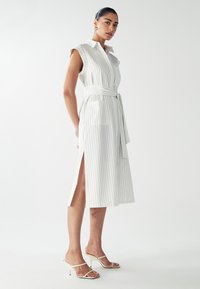 Weißes ärmelloses Kleid mit vertikalen Streifen, Taillengürtel, Seitenschlitzen und Taschen. Das Model kombinierte es mit neutralen Riemchen-Heels.