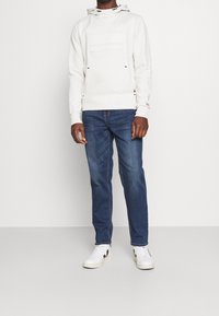 White hooded sweatshirt with a front pocket, textured design, paired with blue denim jeans and white sneakers featuring black accents.