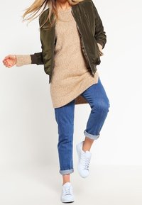 Veste bomber verte olive sur un pull en maille beige, associée à un jean bleu retroussé et des baskets blanches. Textures lisses et coupe décontractée.