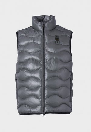 Jeune homme portant un gilet matelassé gris sur un t-shirt noir à manches longues, avec un jean bleu et des baskets noires, debout avec une main dans la poche.