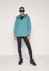 Teal vanntett jakke med glidelås foran og hette, kombinert med svarte leggings med teksturert design og svarte tursko.