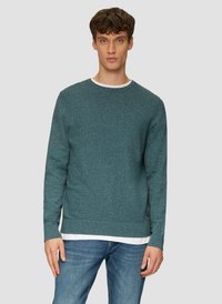 Pull en teal avec un tricot texturé, col rond, et manches longues, porté sur une chemise blanche, associé à un jeans bleu clair.