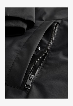 Sort vandtæt jakke stof med et nærbillede af en lynlåslomme med metal lynlås og trykknapdetaljer. Blødt indvendigt krave synlig.