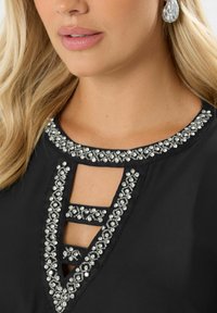 Blusa nera con scollatura a chiave, arricchita con strass d'argento lungo il colletto e dettagli a taglio. Tessuto di trama liscia.