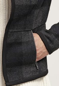 Giacca in misto lana grigia con rifiniture nere, caratterizzata da una tasca con zip sul lato e un tessuto testurizzato con un motivo sottile.