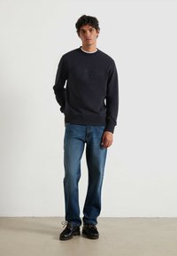 Marineblauwe sweatshirt met verhoogde tekst "FARAH" in donkerblauw, gecombineerd met lichtblauwe recht gesneden jeans en zwarte veterschoenen op een glatte ondergrond.